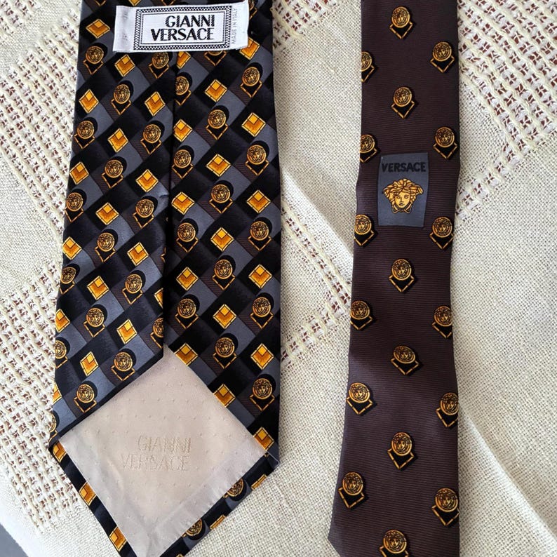 May include: Two silk neckties. One is black with a geometric pattern of gray, gold, and black squares and circles. The other is brown with a repeating gold medallion design. Both ties have the "Gianni Versace" label.