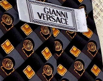 May include: A close-up of a Gianni Versace tie. The tie features a repeating pattern of gold Medusa heads and diamond shapes on a black and gray background. The label reads "GIANNI VERSACE".
