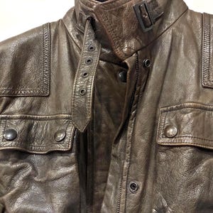May include: A brown leather jacket featuring a textured finish, a high collar with a buckle strap closure, two chest pockets with snap button flaps, and reinforced stitched panels on the shoulders. The jacket includes a front zipper and snap buttons.