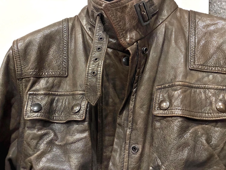 May include: A brown leather jacket featuring a textured finish, a high collar with a buckle strap closure, two chest pockets with snap button flaps, and reinforced stitched panels on the shoulders. The jacket includes a front zipper and snap buttons.