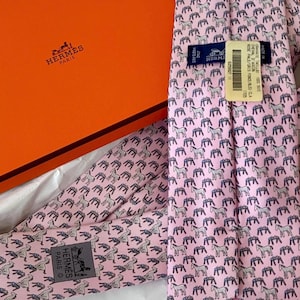 Könnte beinhalten: Eine rosa Seidenkrawatte mit einem sich wiederholenden Muster aus grauen Pferden. Die Krawatte liegt neben einer orangefarbenen Schachtel mit dem Hermes Paris-Logo. Die Krawatte hat ein Etikett mit Text.