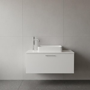 Può includere: Un moderno mobile da bagno bianco con un lavabo rettangolare e un rubinetto cromato. Il mobile ha un singolo cassetto con una maniglia argentata. Lo sfondo è una parete piastrellata grigio chiaro e un pavimento maculato grigio.
