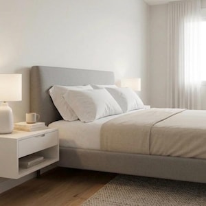 Może przedstawiać: Nowoczesna sypialnia z szarym tapicerowanym łóżkiem, białą pościelą i beżowym kocem. Biała szafka nocna z lampą i małym kubkiem stoi obok łóżka. Pokój ma neutralną paletę kolorów z oknem i białymi zasłonami.