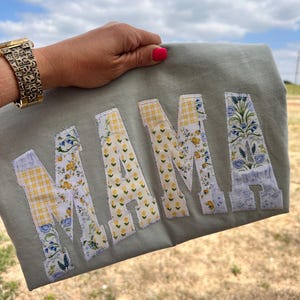 May include: Sage green sweatshirt with the word "MAMA" appliqued in floral fabric. The letters feature yellow, blue, and white floral patterns and yellow gingham. The sweatshirt is held up against a blue sky.