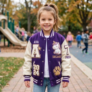 May include: A purple and cream varsity jacket featuring the name AVA, the number 7, the year 2024, a unicorn graphic with the text DREAMER, the word PRIDE, and a paw print patch. The jacket has snap buttons and striped ribbed cuffs and collar.