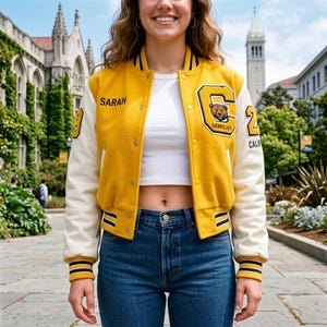 Könnte beinhalten: Eine gelb-cremefarbene Collegejacke mit dem Namen SARAH auf der rechten Brust, einem großen Buchstaben C mit Bärengrafik und dem Schriftzug BERKELEY auf der linken Brust sowie der Zahl 23 und dem Schriftzug CALIF auf den Ärmeln.