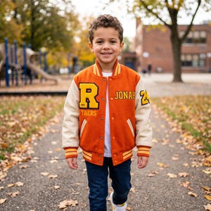 May include: A varsity jacket featuring a burnt orange wool body with cream-colored sleeves. The front displays a large yellow letter R with the word TIGERS below it, the name JONAH, and the number 24 on the sleeve, all with yellow and orange trim.