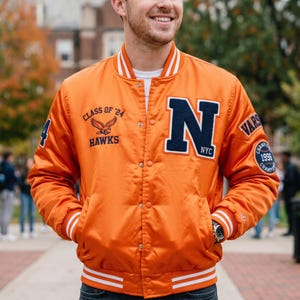 Puede incluir: Una chaqueta universitaria naranja con puños y cuello de canalé a rayas blancas. El frente presenta una gran letra N azul marino, el texto Class of '24 Hawks con un gráfico de águila y un parche en la manga 1996 Champion.