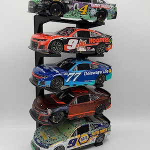 May include: A vertical black display stand holding five die-cast stock car models. The cars feature various colorful liveries, including Grave Digger, Hooters, Delaware Life, and NAPA Auto Parts, with detailed racing decals and black wheels.