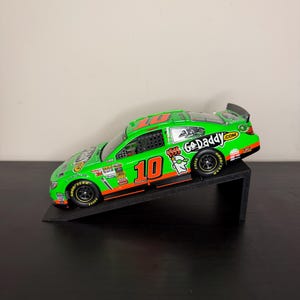 Può includere: Un modello di auto da corsa verde e arancione con il numero 10, su un supporto nero. L'auto presenta il logo GoDaddy.com e pneumatici Goodyear. L'auto è inclinata verso l'alto.