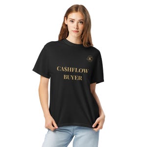 May include: A black short-sleeve t-shirt featuring the gold text CA$HFLOW BUYER printed on the chest. A small circular gold logo with the letter K is positioned above the text. The shirt has a crew neckline and a standard fit design.