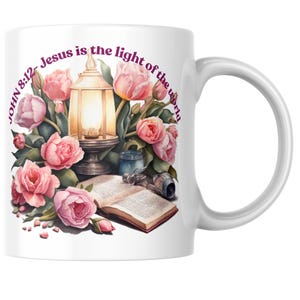 May include: White ceramic mug with a watercolour design of pink roses, tulips, and a lit lantern. The text "John 8:12 - Jesus is the light of the world" is printed in a curved font around the floral arrangement. A book and other objects are also depicted.