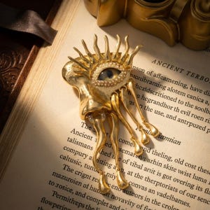 Op de afbeelding: Goudkleurige broche in de vorm van een oog met een blauwe iris, omringd door parels en zonnestralen. De broche heeft bungelende poten en ligt op een boek met de tekst "ANCIENT TERBO."