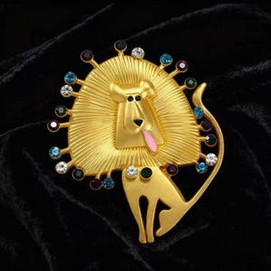 Könnte beinhalten: Goldfarbene Löwenbrosche mit strukturierter Mähne und Körper. Der Löwe hat eine rosa Zunge herausgestreckt und ist mit bunten Edelsteinen in verschiedenen Farben, darunter Blau, Grün und Klar, verziert. Die Brosche ist vor einem schwarzen Hintergrund platziert.
