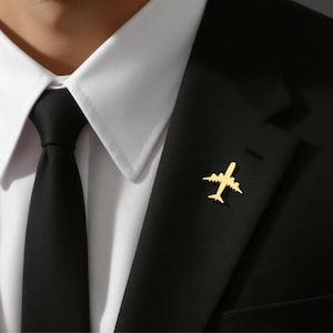 May include: A gold-coloured airplane lapel pin on a black suit jacket. The pin is a detailed silhouette of an airplane. The jacket is worn over a white dress shirt and a black tie.