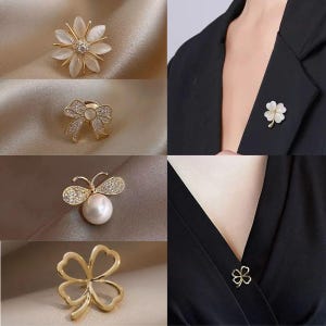 Puede incluir: Una colección de broches dorados. Los broches incluyen una flor con pétalos blancos, un lazo con cristales transparentes, una abeja con cuerpo de perla y un trébol de cuatro hojas. Dos broches se muestran sobre ropa negra.