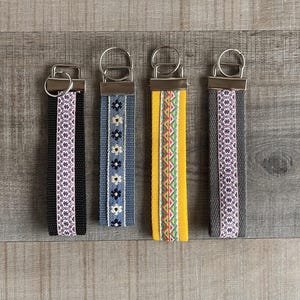 Könnte beinhalten: Vier Schlüsselanhänger mit dekorativen Stoffbändern. Die Bänder weisen verschiedene Muster auf, darunter Blumen- und geometrische Designs, in Farben wie Schwarz, Blau, Gelb und Grau. Jeder Schlüsselanhänger hat einen silberfarbenen Metallring.