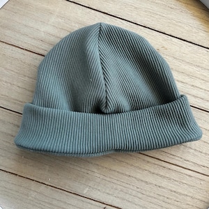 Könnte beinhalten: Eine einfarbige, salbeigrüne Beanie-Mütze mit durchgehender Rippstrickstruktur. Die Mütze ist mit einem breiten, umgeschlagenen Saum am unteren Rand gestaltet, was für eine strukturierte und klassische Silhouette sorgt. Das Material wirkt weich und einheitlich.
