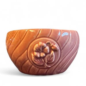 Könnte beinhalten: Eine braune Keramikschale mit floralem Design. Die Schale hat eine breite, abgerundete Form und eine strukturierte Oberfläche mit einem geschnitzten Blumenmedaillon. Die Schale hat einen Durchmesser von etwa 15 cm.