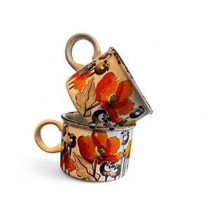 May include: Two ceramic mugs stacked on top of each other. The mugs are white with a floral design featuring orange and black flowers. Each mug has a rounded handle.