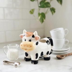 May include: A ceramic cow-shaped creamer pitcher featuring a white body with black spots, a light tan muzzle, and pink-tinted ears. The pitcher has a small opening at the mouth for pouring and a black tail handle, standing on four black hooves.