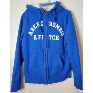 May include: A blue zip-up hoodie with the text "Abercrombie & Fitch" in white. The hoodie has a fleece-lined hood and two front pockets. The cuffs and hem are ribbed.
