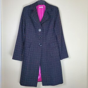 May include: A long-sleeved coat in a dark charcoal tweed fabric with a subtle grid pattern and flecks of orange and yellow. The coat features a notched lapel, three black buttons down the front, two flap pockets, and a vibrant hot pink lining.