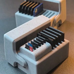 May include: A light gray speckled storage case with a hinged lid, designed to hold multiple SD and microSD memory cards in vertical slots. The case features a textured finish and contains several black memory cards and one blue card.