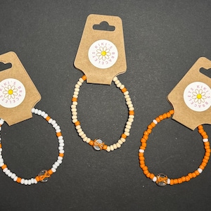 May include: Three beaded bracelets on brown tags. One bracelet is white and orange, another is cream and orange, and the third is orange. Each bracelet has a silver-colored bead. The tags have a daisy logo and the text "Kiki's Bracelets".
