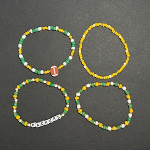 May include: Four beaded bracelets in green, yellow, white, and gold. One bracelet features a football charm, and another spells "PACKERS" with letter beads. The bracelets are on a black surface.