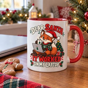 Op de afbeelding: Een witte keramische mok met een rode binnenkant en handvat, voorzien van een afbeelding van een vos met een kerstmuts en een groene trui met patroon, zittend met een laptop en een mok. De tekst luidt: Dear Santa It Worked On My Laptop.
