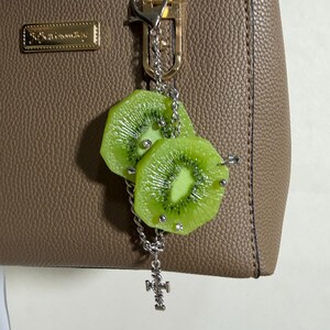 May include: A bag charm featuring two realistic green kiwi fruit slices attached to a silver-toned metal chain. The kiwi slices are accented with small silver studs and clear rhinestones. A small silver-toned metal cross charm hangs from the bottom of the chain, which is connected to a silver lobster clasp.