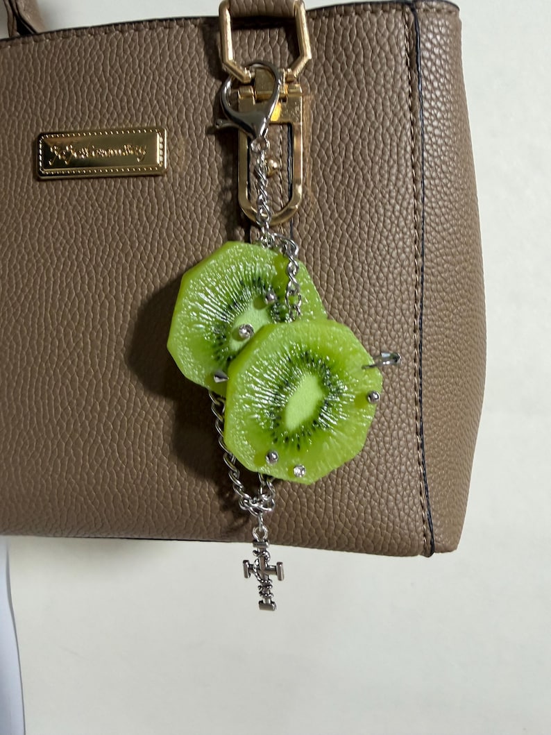 May include: A bag charm featuring two realistic green kiwi fruit slices attached to a silver-toned metal chain. The kiwi slices are accented with small silver studs and clear rhinestones. A small silver-toned metal cross charm hangs from the bottom of the chain, which is connected to a silver lobster clasp.