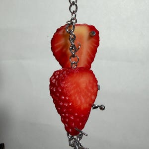 May include: A silver metal chain keychain featuring two strawberry halves attached with silver metal piercing jewelry. The strawberries are bright red with visible seeds and white centers, connected by a vertical chain and small metal barbell studs.