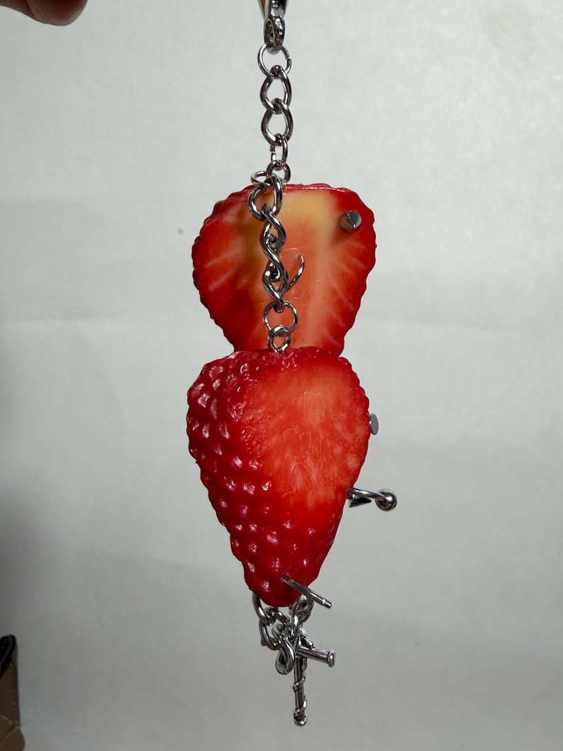 May include: A silver metal chain keychain featuring two strawberry halves attached with silver metal piercing jewelry. The strawberries are bright red with visible seeds and white centers, connected by a vertical chain and small metal barbell studs.