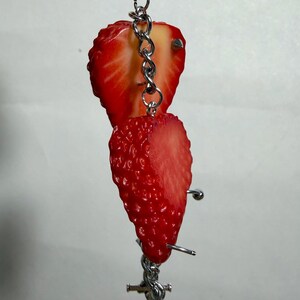 May include: A silver-toned metal chain keychain featuring two realistic strawberry slices. The bright red fruit pieces are attached to the vertical chain, which includes small metal studs and a toggle clasp at the bottom, creating a dangling accessory.