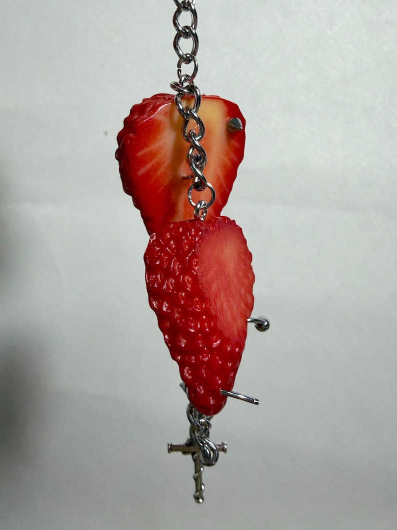 May include: A silver-toned metal chain keychain featuring two realistic strawberry slices. The bright red fruit pieces are attached to the vertical chain, which includes small metal studs and a toggle clasp at the bottom, creating a dangling accessory.