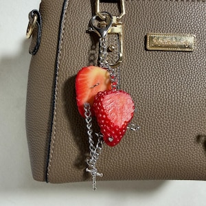 Peut inclure: Un sac à main texturé taupe avec une plaque logo en métal doré et un porte-clés argenté. Le porte-clés se compose de deux tranches de fraise réalistes et d'une petite croix argentée fixées à une chaîne en métal argenté avec un mousqueton.