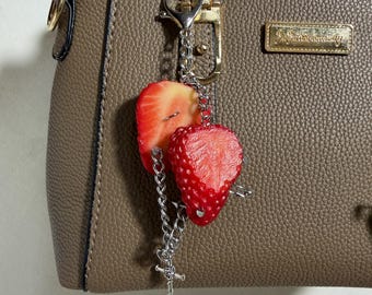 May include: A taupe textured handbag with a gold-toned metal logo plate and a silver-toned keychain accessory. The keychain features two realistic strawberry slices and a small silver cross charm attached to a silver metal chain and lobster clasp.