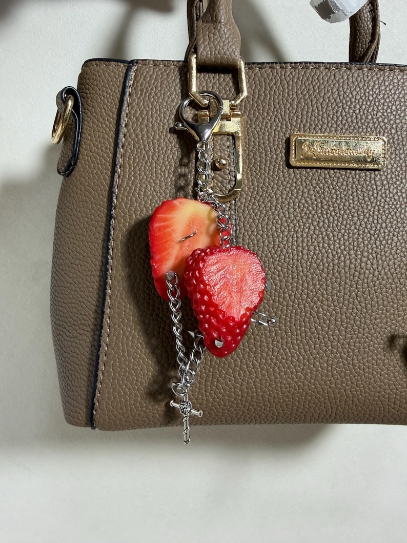 May include: A taupe textured handbag with a gold-toned metal logo plate and a silver-toned keychain accessory. The keychain features two realistic strawberry slices and a small silver cross charm attached to a silver metal chain and lobster clasp.