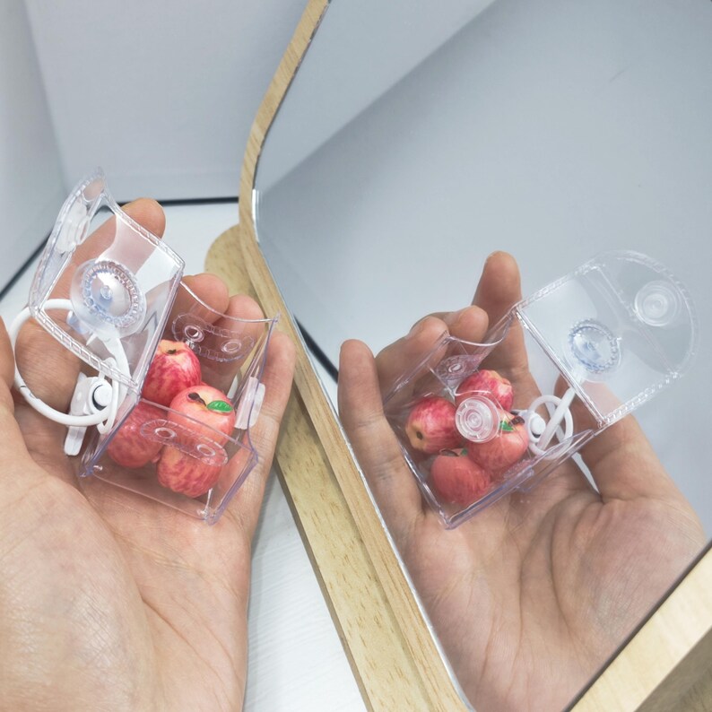 May include: Clear plastic container with a hinged lid, holding miniature red apples. The container has a suction cup and a white loop. The image is reflected in a mirror.
