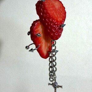 May include: A decorative hanging ornament featuring two bright red strawberry slices pierced by silver-toned metal spikes and curved barbell jewelry. A silver-toned metal chain hangs from the fruit, ending in a small, detailed sword-shaped charm.