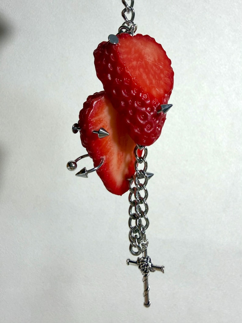 May include: A decorative hanging ornament featuring two bright red strawberry slices pierced by silver-toned metal spikes and curved barbell jewelry. A silver-toned metal chain hangs from the fruit, ending in a small, detailed sword-shaped charm.