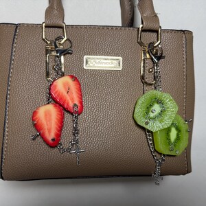 May include: A taupe textured handbag featuring two silver-toned metal chains attached to the handles. One chain holds two red strawberry slices, and the other chain holds two green kiwi slices. A small rectangular gold-toned plaque is centered on the bag.
