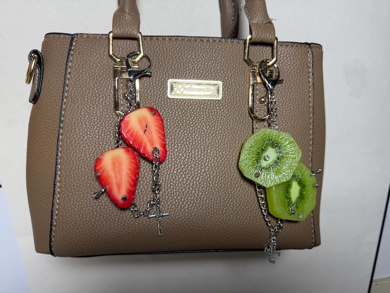 May include: A taupe textured handbag featuring two silver-toned metal chains attached to the handles. One chain holds two red strawberry slices, and the other chain holds two green kiwi slices. A small rectangular gold-toned plaque is centered on the bag.