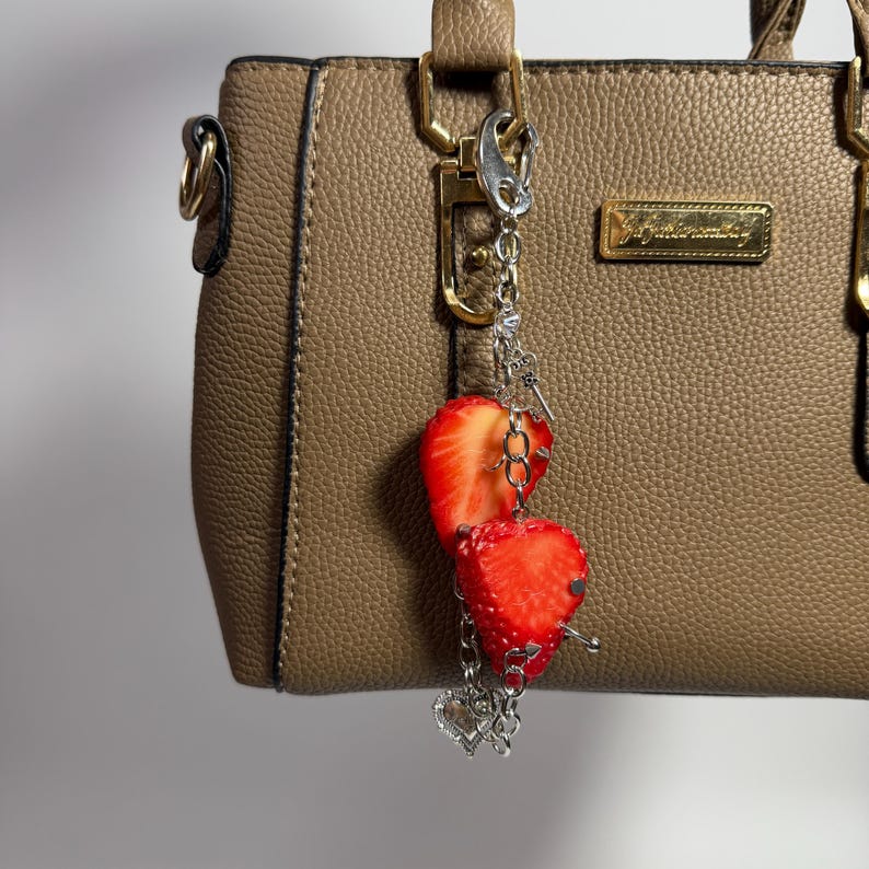May include: A taupe textured handbag with a gold-toned rectangular logo plate and a silver-toned metal bag charm. The charm features two realistic strawberry slices, a small silver key, and a dangling heart-shaped pendant on a link chain.