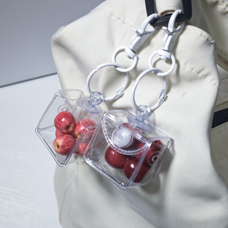 May include: Two clear, square containers with white metal clasps and rings, each holding small red apples. The containers are attached to a white jacket. The apples have a red and yellow colour.