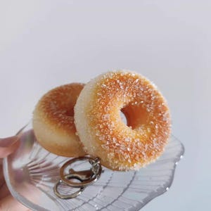 May include: Two sugared donuts on a clear, seashell-shaped plate with a silver keychain. The donuts are golden brown with a generous coating of coarse sugar crystals. The plate is held by a hand, with a plain white background.