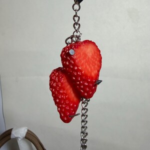 May include: A keychain featuring two realistic red strawberry halves attached to a silver-toned metal chain. The strawberries are pierced with small silver metal studs and spikes, connected to a circular metal ring clasp at the top.