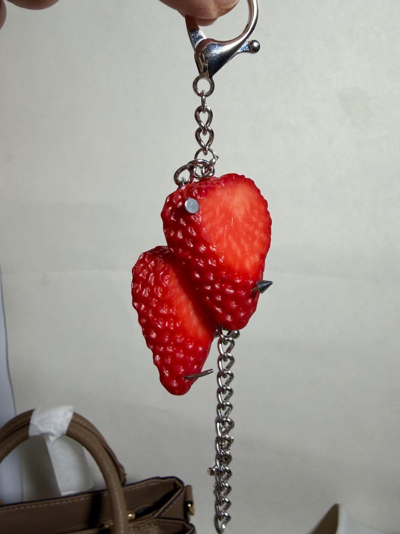 May include: A keychain featuring two realistic red strawberry halves attached to a silver-toned metal chain. The strawberries are pierced with small silver metal studs and spikes, connected to a circular metal ring clasp at the top.